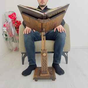 K&ouml;nnte beinhalten: Ein B&uuml;cherst&auml;nder aus Holz mit aufwendigen Schnitzereien. Der St&auml;nder ist verstellbar und hat ein braunes Lederbuch darauf liegen. Das Buch tr&auml;gt den Titel "Maltepe" in goldenen Lettern.