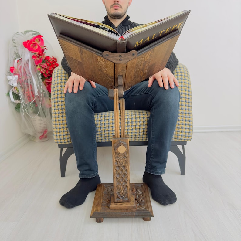 K&ouml;nnte beinhalten: Ein B&uuml;cherst&auml;nder aus Holz mit aufwendigen Schnitzereien. Der St&auml;nder ist verstellbar und hat ein braunes Lederbuch darauf liegen. Das Buch tr&auml;gt den Titel "Maltepe" in goldenen Lettern.