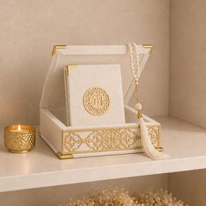 Puede incluir: Una caja de terciopelo color crema con esquinas de metal dorado y una banda decorativa de filigrana dorada. Dentro, un libro color crema a juego presenta un emblema circular dorado con caligrafía árabe. Un rosario de perlas blancas con borla.