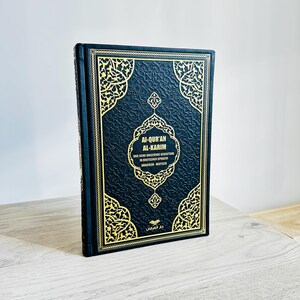 May include: A hardcover book with a black cover and gold filigree design. The title "Al-Qur'an Al-Karim" is displayed in white text, with additional text in German and Arabic. The book is standing upright on a light-colored surface.