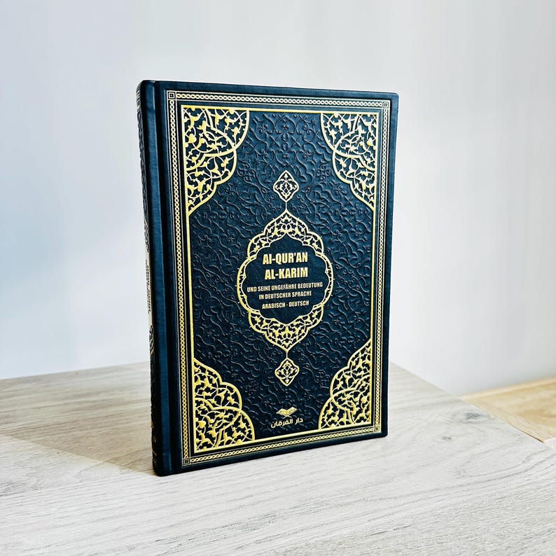 May include: A hardcover book with a black cover and gold filigree design. The title "Al-Qur'an Al-Karim" is displayed in white text, with additional text in German and Arabic. The book is standing upright on a light-colored surface.