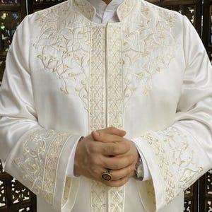 May include: White long-sleeved robe with intricate gold embroidery on the front and cuffs. The embroidery features a floral pattern.