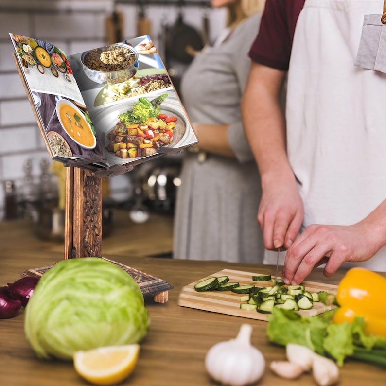 K&ouml;nnte beinhalten: Ein Holz-Kochbuchst&auml;nder mit einem Kochbuch, das auf einer Seite mit verschiedenen Rezepten ge&ouml;ffnet ist. Der St&auml;nder steht auf einem Holztisch mit verschiedenen frischen Gem&uuml;sesorten und einem Messer.