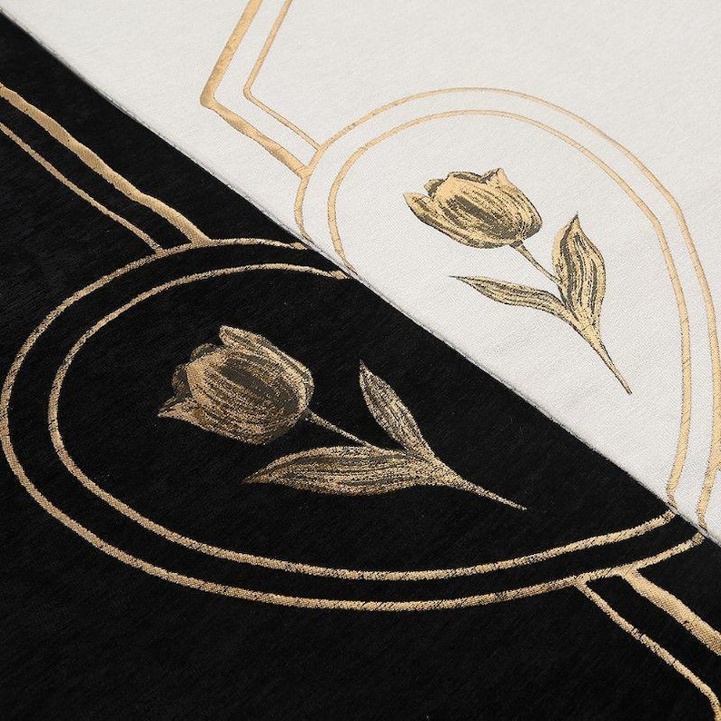 May include: Close-up of a black and white textile with gold floral designs. The design features a tulip in an oval frame, with gold lines and accents. The background is split diagonally, with black on the bottom left and white on the top right.