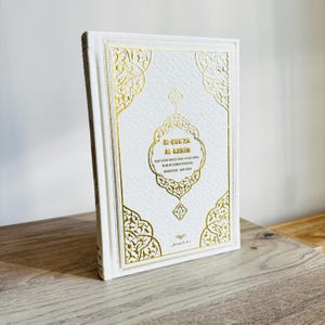 May include: A white book with gold decorative accents and text. The cover features ornate gold filigree designs and the title "AL-QUR'AN AL-KARIM" in gold lettering. The book is standing upright on a wooden surface.