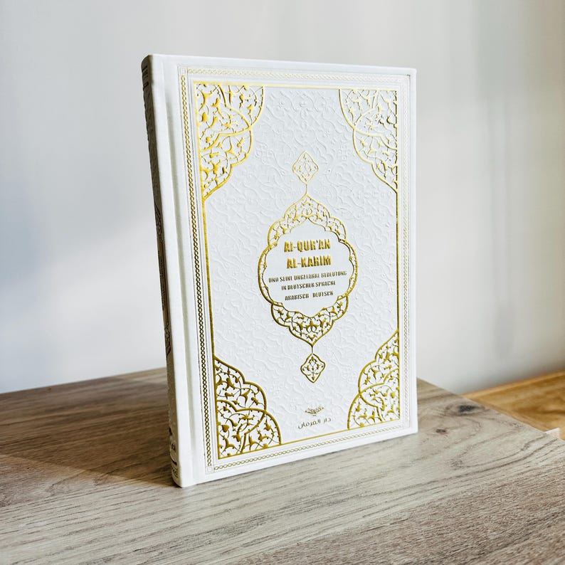 May include: A white book with gold decorative accents and text. The cover features ornate gold filigree designs and the title "AL-QUR'AN AL-KARIM" in gold lettering. The book is standing upright on a wooden surface.