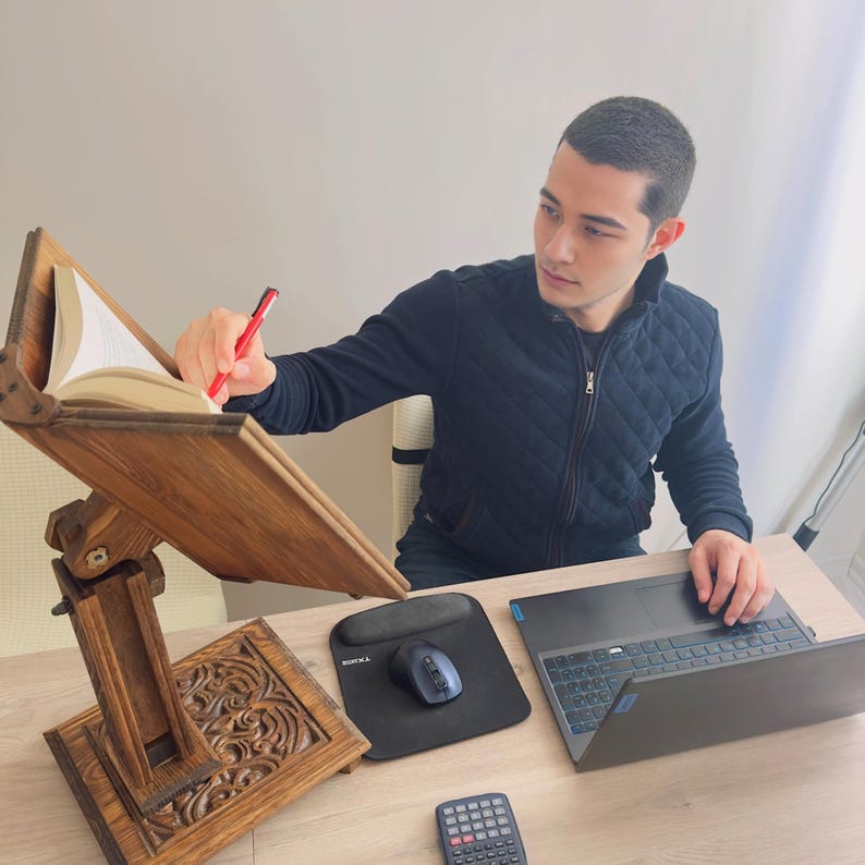 K&ouml;nnte beinhalten: Ein h&ouml;lzerner Buchst&auml;nder mit aufwendigen Schnitzereien, der ein offenes Buch h&auml;lt. Eine Person sitzt an einem Schreibtisch mit einem Laptop und einem Mauspad und verwendet einen roten Stift, um auf das Buch zu zeigen.