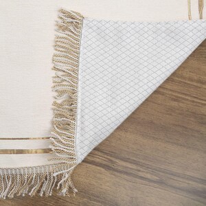 May include: Cream-colored rug with a woven design and gold stripes. The rug has a fringe border in shades of beige and cream. The underside of the rug is visible, showing a white diamond pattern. The rug is partially rolled up, revealing the underside.