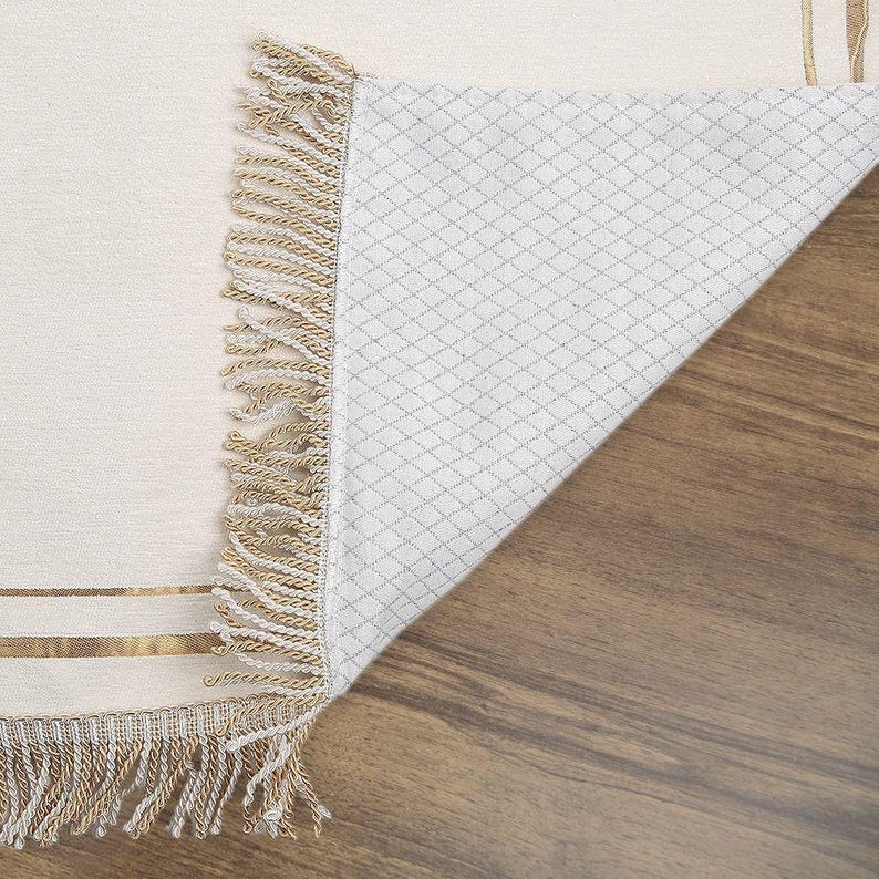May include: Cream-colored rug with a woven design and gold stripes. The rug has a fringe border in shades of beige and cream. The underside of the rug is visible, showing a white diamond pattern. The rug is partially rolled up, revealing the underside.