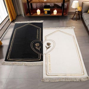 May include: Two prayer rugs, one black and one white, with gold trim and fringe. Each rug features a gold floral design and the text "CANTIM BABAM" and "CANTIM ANNEM". The rugs are rectangular with an arched top.