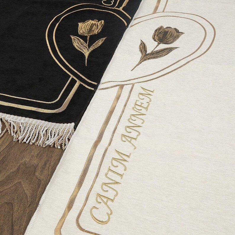 May include: Two prayer rugs, one black and one cream, with gold floral embroidery. Each rug features a tulip design within a heart-shaped border and the text "CANIM ANNEM" in gold.