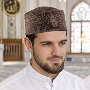 May include: A brown velvet kufi hat with detailed embroidery. The hat has a square crown and an intricate pattern. The person in the image is wearing a white shirt. The background is a mosque.