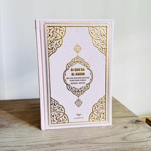 May include: A pink hardcover book titled "Al-Qur'an Al-Karim" with gold decorative accents. The book's cover features ornate gold patterns and text in both Arabic and German. The book is standing upright on a wooden surface.