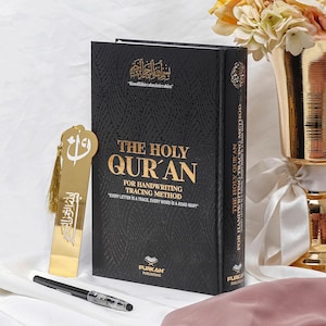 May include: A black hardback book with gold lettering titled "The Holy Qur'an" for handwriting tracing method. The book has the text "Every letter is a trace, every word is a road map" printed on the cover. The book is next to a gold bookmark with an Islamic symbol.