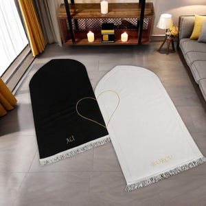 Puede incluir: Dos alfombras de oración, una negra y otra blanca, se muestran sobre un suelo embaldosado. La alfombra negra tiene el nombre "ALI" bordado en dorado, mientras que la alfombra blanca tiene "BURCU". Ambas alfombras tienen un borde con flecos y están conectadas por un diseño dorado en forma de corazón.
