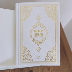 以下が含まれることがあります： 金色の装飾が施された白い本で、タイトルは「THE HOLY QURAN ARABIC - ENGLISH」と金色の文字で書かれています。表紙にはエンボス加工の模様と豪華な金の縁取りが施されています。本は開かれており、白いページが見えています。
