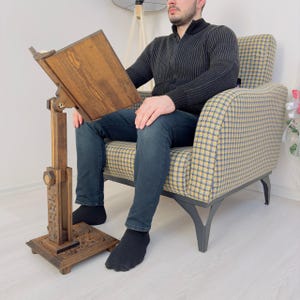 K&ouml;nnte beinhalten: Ein B&uuml;cherst&auml;nder aus Holz mit einem geschnitzten Sockel und einstellbarer H&ouml;he. Der St&auml;nder ist aus dunkelbraunem Holz gefertigt und hat einen rechteckigen Sockel. Der St&auml;nder wird von einer Person verwendet, die auf einem Stuhl sitzt.