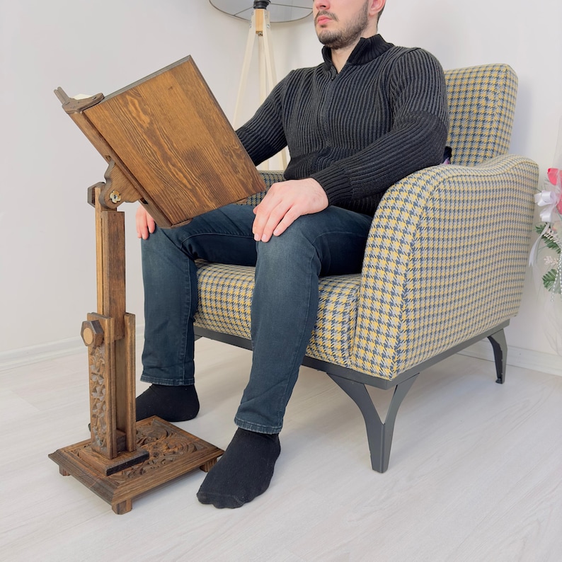 K&ouml;nnte beinhalten: Ein B&uuml;cherst&auml;nder aus Holz mit einem geschnitzten Sockel und einstellbarer H&ouml;he. Der St&auml;nder ist aus dunkelbraunem Holz gefertigt und hat einen rechteckigen Sockel. Der St&auml;nder wird von einer Person verwendet, die auf einem Stuhl sitzt.