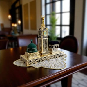 May include: A gold-colored decorative set featuring a clock tower, a dome, and a small black cube, likely representing religious landmarks. The set is placed on a decorative, lace-edged mat on a dark wooden table.