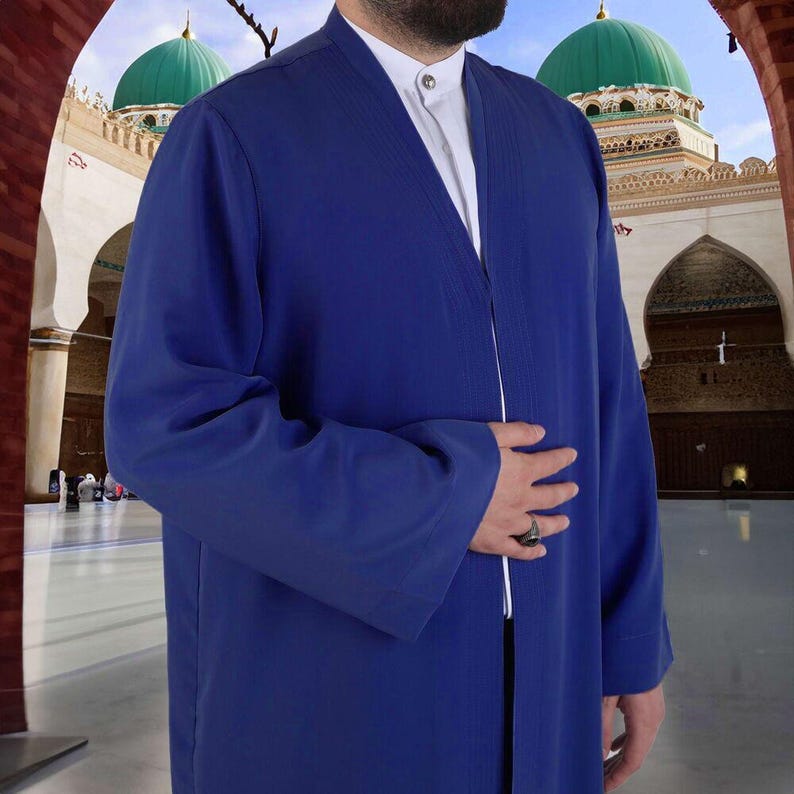 Op de afbeelding: Een lange, koningsblauwe jurk met wijde mouwen. De jurk wordt gedragen over een wit overhemd met kraag. De achtergrond toont een moskee met groene koepels en gewelfde ingangen. De jurk heeft een eenvoudig, elegant ontwerp.
