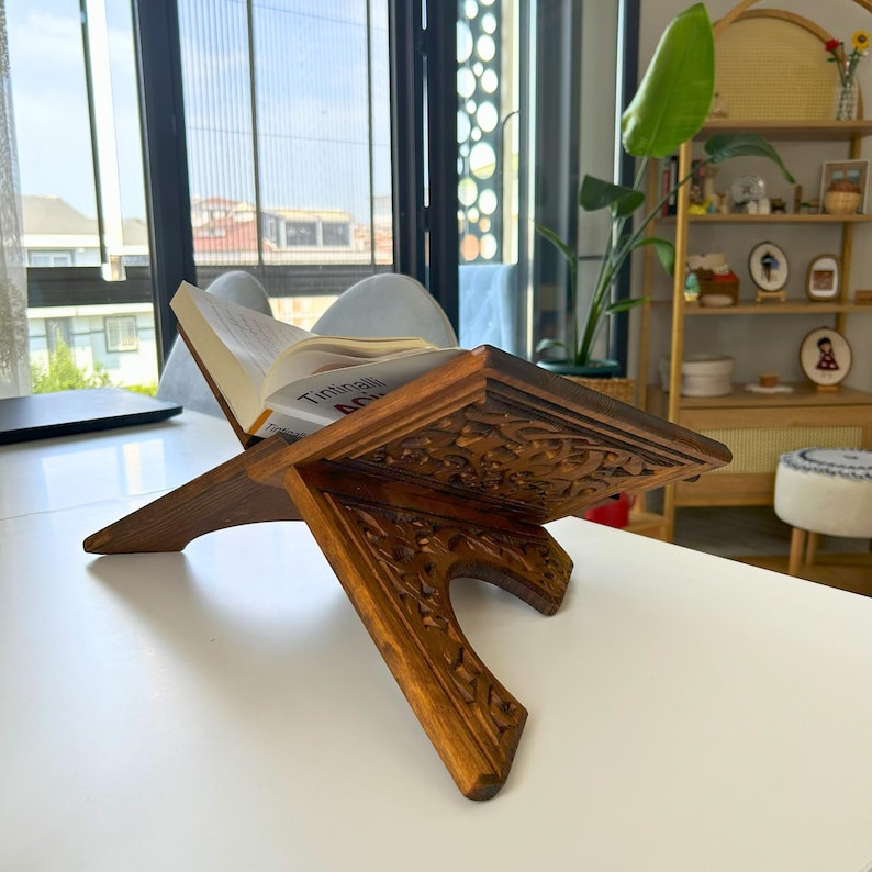 Puede incluir: Un atril de madera tallada, mostrando un libro abierto. La madera es de color marr&oacute;n c&aacute;lido y presenta intrincados dise&ntilde;os florales. El atril est&aacute; colocado sobre una superficie blanca, con una ventana y decoraci&oacute;n interior en el fondo.