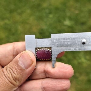 Peut inclure: Une bague rectangulaire en rubis est mesur&eacute;e avec un pied &agrave; coulisse. Le rubis est d'un rouge profond, serti dans une monture en m&eacute;tal dor&eacute;. Le pied &agrave; coulisse indique que la pierre mesure environ 2 cm de long.
