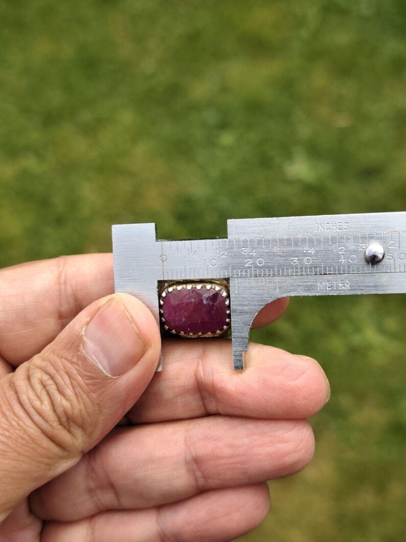 Peut inclure: Une bague rectangulaire en rubis est mesur&eacute;e avec un pied &agrave; coulisse. Le rubis est d'un rouge profond, serti dans une monture en m&eacute;tal dor&eacute;. Le pied &agrave; coulisse indique que la pierre mesure environ 2 cm de long.