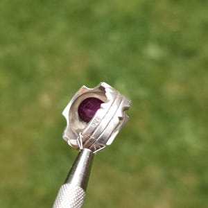 Peut inclure: Une bague en argent orn&eacute;e d'une pierre pr&eacute;cieuse violet fonc&eacute;. La bague pr&eacute;sente un design textur&eacute; unique avec une s&eacute;rie de petits bords pointus sur le dessus. La bague est tenue par un outil argent&eacute; sur un fond vert flou.