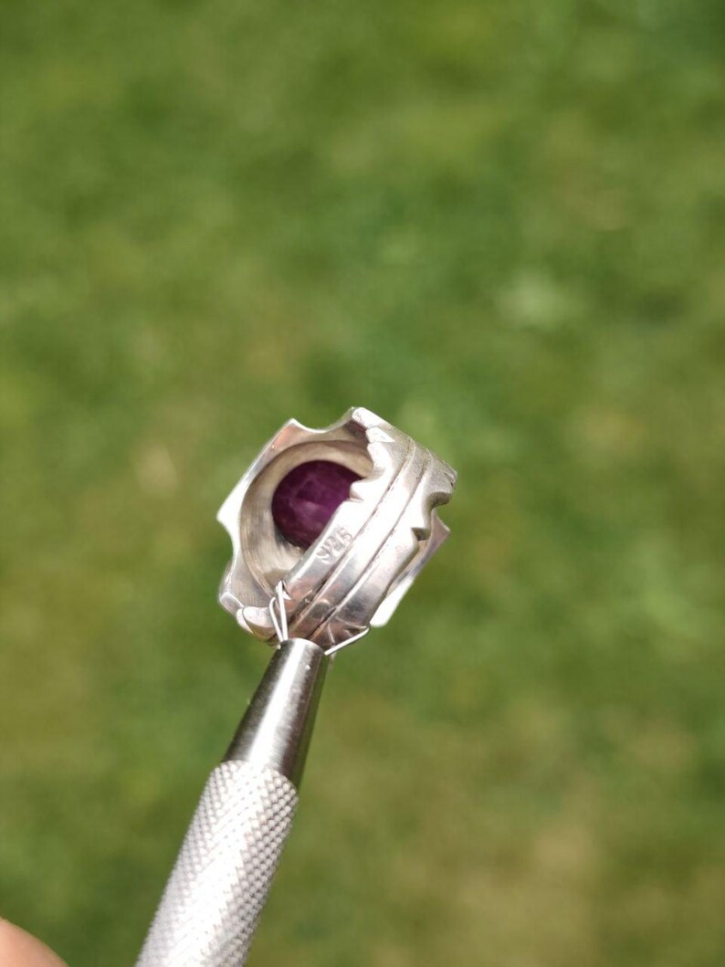 Peut inclure: Une bague en argent orn&eacute;e d'une pierre pr&eacute;cieuse violet fonc&eacute;. La bague pr&eacute;sente un design textur&eacute; unique avec une s&eacute;rie de petits bords pointus sur le dessus. La bague est tenue par un outil argent&eacute; sur un fond vert flou.