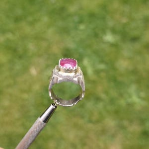 Peut inclure: Une bague en argent orn&eacute;e d'un grand rubis taill&eacute; en carr&eacute;. La bague est tenue par un outil argent&eacute; sur un fond vert flou. Le rubis est d'un rose profond et est serti dans une monture argent&eacute;e d&eacute;corative.