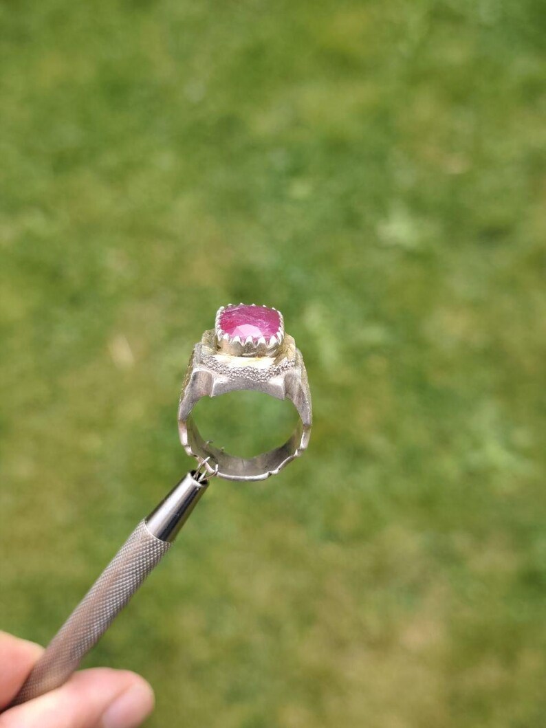 Peut inclure: Une bague en argent orn&eacute;e d'un grand rubis taill&eacute; en carr&eacute;. La bague est tenue par un outil argent&eacute; sur un fond vert flou. Le rubis est d'un rose profond et est serti dans une monture argent&eacute;e d&eacute;corative.