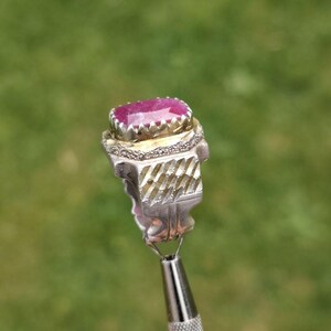 Peut inclure: Une bague en argent orn&eacute;e d'une grosse pierre rose ovale. La bague pr&eacute;sente un motif textur&eacute; et d&eacute;taill&eacute; autour de la pierre et sur le corps. La bague est tenue par un outil argent&eacute; sur un fond vert flou.
