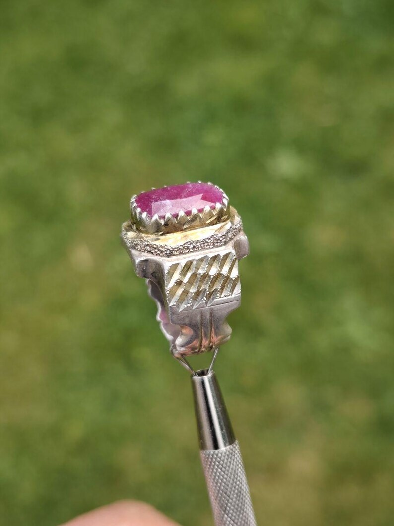 Peut inclure: Une bague en argent orn&eacute;e d'une grosse pierre rose ovale. La bague pr&eacute;sente un motif textur&eacute; et d&eacute;taill&eacute; autour de la pierre et sur le corps. La bague est tenue par un outil argent&eacute; sur un fond vert flou.