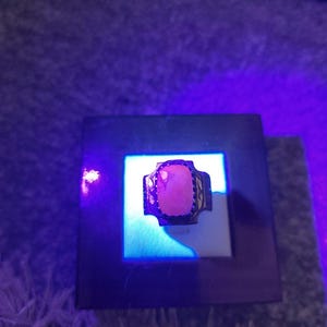 Peut inclure: Une bague en argent avec une grosse pierre rose rectangulaire. La bague est pr&eacute;sent&eacute;e sur une surface blanche dans un cadre carr&eacute; fonc&eacute;, &eacute;clair&eacute;e par une lumi&egrave;re bleue. Le sertissage de la bague pr&eacute;sente un motif d&eacute;coratif.