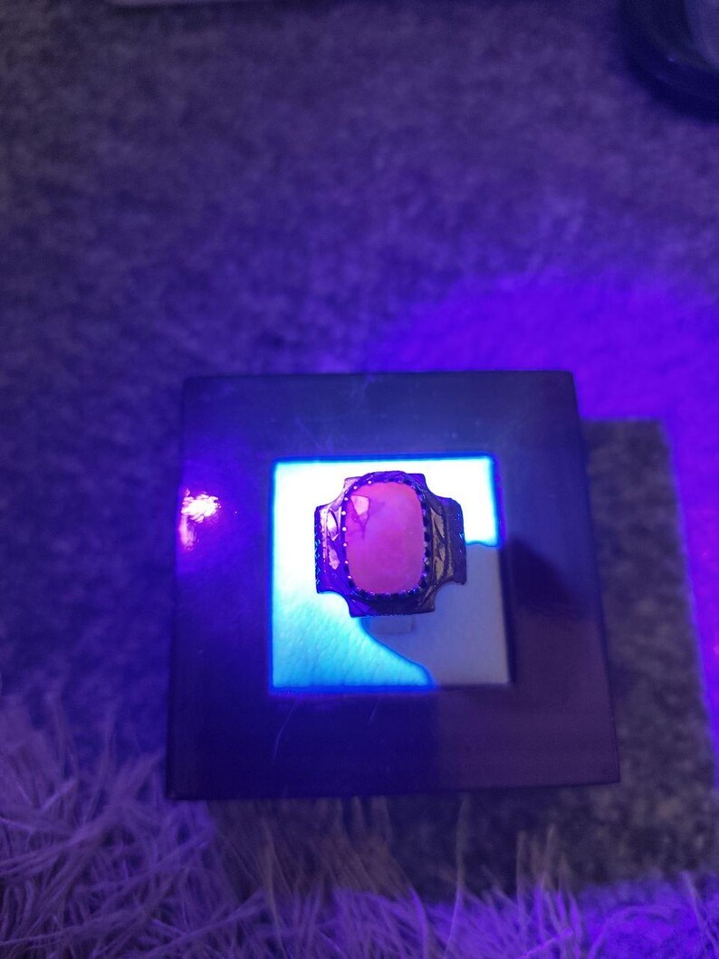 Peut inclure: Une bague en argent avec une grosse pierre rose rectangulaire. La bague est pr&eacute;sent&eacute;e sur une surface blanche dans un cadre carr&eacute; fonc&eacute;, &eacute;clair&eacute;e par une lumi&egrave;re bleue. Le sertissage de la bague pr&eacute;sente un motif d&eacute;coratif.