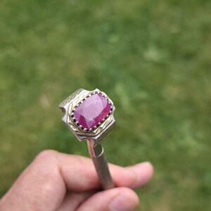 Peut inclure: Une bague argent&eacute;e orn&eacute;e d'une grosse pierre pr&eacute;cieuse rectangulaire rouge fonc&eacute;. La pierre est sertie dans une lunette d&eacute;corative avec une bordure dor&eacute;e. La bague est tenue sur un fond vert flou.