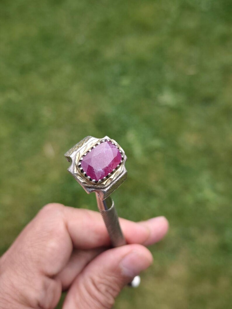 Peut inclure: Une bague argent&eacute;e orn&eacute;e d'une grosse pierre pr&eacute;cieuse rectangulaire rouge fonc&eacute;. La pierre est sertie dans une lunette d&eacute;corative avec une bordure dor&eacute;e. La bague est tenue sur un fond vert flou.