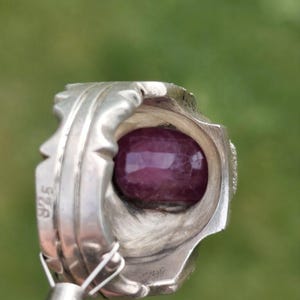 Peut inclure: Bague en argent avec une grosse pierre pr&eacute;cieuse ovale, rouge fonc&eacute;. La bague pr&eacute;sente un design orn&eacute; et d&eacute;taill&eacute; avec une bande textur&eacute;e et le chiffre "925" estampill&eacute; sur le c&ocirc;t&eacute;. La pierre pr&eacute;cieuse polie refl&egrave;te la lumi&egrave;re.