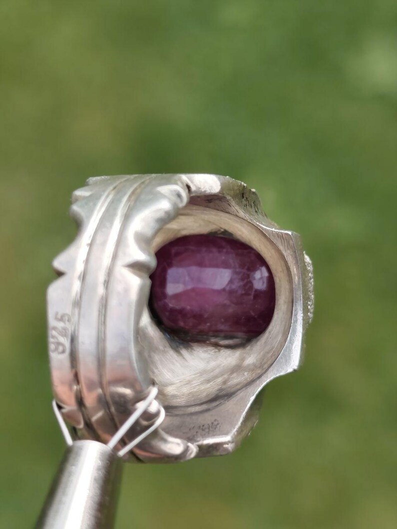 Peut inclure: Bague en argent avec une grosse pierre pr&eacute;cieuse ovale, rouge fonc&eacute;. La bague pr&eacute;sente un design orn&eacute; et d&eacute;taill&eacute; avec une bande textur&eacute;e et le chiffre "925" estampill&eacute; sur le c&ocirc;t&eacute;. La pierre pr&eacute;cieuse polie refl&egrave;te la lumi&egrave;re.