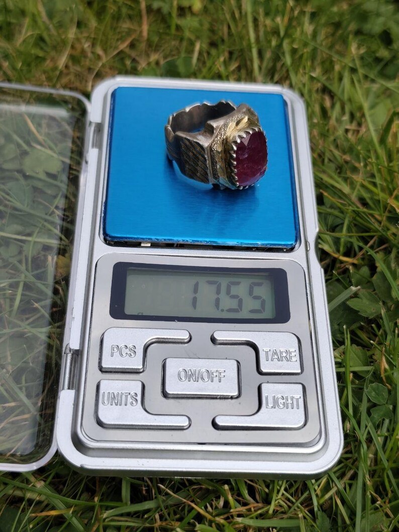Peut inclure: Une bague argent&eacute;e avec une grosse pierre pr&eacute;cieuse ovale rouge fonc&eacute; est pr&eacute;sent&eacute;e sur une balance num&eacute;rique. La balance affiche un poids de 17,55. La bague a un design orn&eacute; et textur&eacute;.