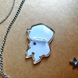 Op de afbeelding: Een heldere acrylspeld met een witte achterkant en een veiligheidsspeldsluiting. De speld heeft een cartoon-personageontwerp. Een zilveren ketting en een stervormige bedel zijn zichtbaar op de achtergrond.