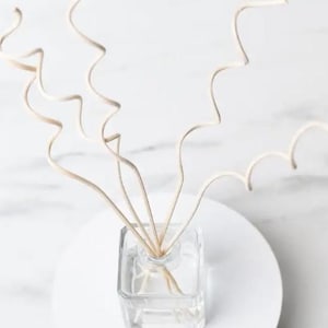 May include: A clear, square glass diffuser bottle with light-colored reeds. The bottle sits on a white, round surface. The reeds have a curly, wavy design, adding a decorative element to the home fragrance product.