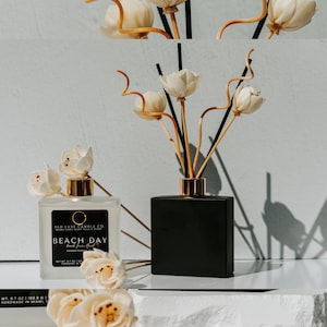 May include: A black square glass diffuser bottle with a gold lid and wooden diffuser sticks. The bottle is sitting on a white surface with white flowers. The bottle has a label that says "ELO LUXE CANDLE CO" and "BEACH DAY".