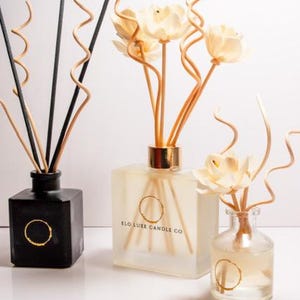 May include: Three decorative reed diffusers in varying sizes. The diffusers are in black, frosted, and clear glass bottles. The bottles contain reeds and decorative flower accents. The frosted bottle has the text "ELO LUXE CANDLE CO" and a gold circle logo.