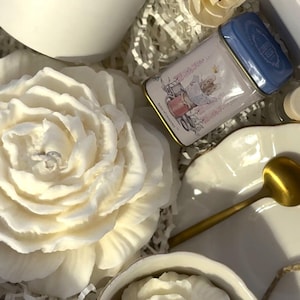 Op de afbeelding: Een geschenkdoos met een witte kaars, een kleine blikken doos met een blauw deksel en een witte roosvormige kaars. De doos bevat ook een kleine witte theekop met een roosvormige kaars, een gouden lepel en een wit bord.