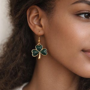 May include: Gold-tone shamrock earrings with a dark green enamel finish. The earrings feature a three-leaf clover design, with a gold hook for pierced ears. The earrings are a classic accessory for St. Patrick's Day or everyday wear.