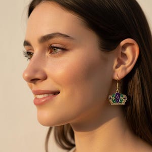 May include: Gold-tone crown earrings with purple, green, and clear accents. The earrings feature a detailed crown design with a hook closure. The earrings are a festive accessory.