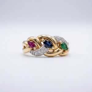 Pode incluir: Um anel de ouro amarelo com design trançado e três pedras preciosas de corte marquise: um rubi vermelho, uma safira azul e uma esmeralda verde. A banda é acentuada com pequenos diamantes redondos transparentes cravados em pavé.