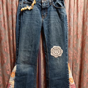 May include: Blue denim jeans with decorative elements. Features include a crocheted patch, floral fabric inserts, and a yellow crocheted trim on the pocket. The jeans are hanging on a hanger.