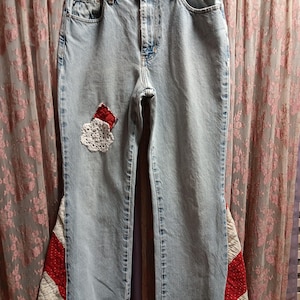 May include: Light blue denim jeans with a high waist and a straight leg. The jeans feature a decorative patch on the front and a patchwork design in red, white, and purple on the outer seams. The jeans are hanging on a white hanger.